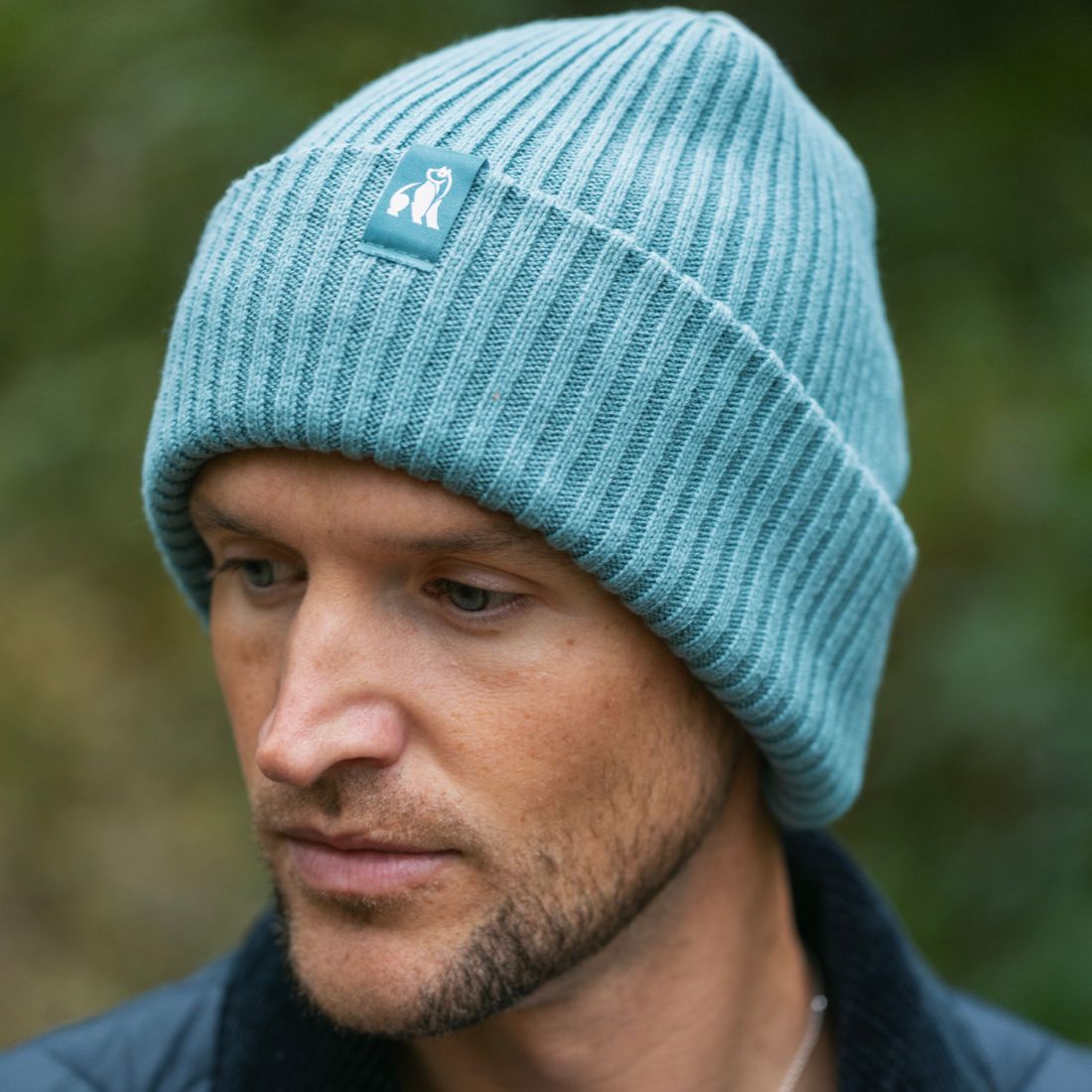 Man wearing a light blue knit beanie with a logo against a blurred natural background