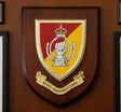 A wooden shield-shaped plaque featuring the Royal Armoured Corps (RAC) crest, mounted on a brown wood-paneled wall. The crest has a red and yellow background with a white raised fist under a crown, and a gold scroll reading 'ROYAL ARMOURED CORPS'.