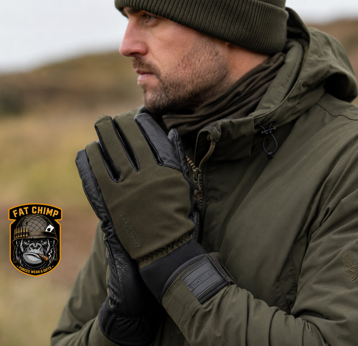 A man wearing an olive green hooded jacket, matching beanie, and black-and-olive gloves stands outdoors in a blurred natural setting. A 'Fat Chimp' brand patch is visible in the lower-left corner.