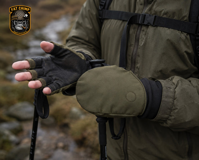 A close-up of a person outdoors wearing olive green insulated gloves with black cuffs and palm areas, paired with an olive green jacket and backpack straps. A 'Fat Chimp' brand logo is visible in the top-left corner.