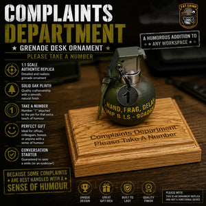 A humorous desk ornament shaped like a military grenade with printed text reading 'COMPLAINTS DEPARTMENT' and 'Please Take A Number,' displayed on a wooden box with product feature callouts around it.