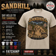 A beige graphic t-shirt displayed on a dark promotional background. The shirt features a large vintage-style military-themed graphic with the text 'Pirbright Sandhill' and 'Say Hello to the Sandhill' with a photo of soldiers on a trail.