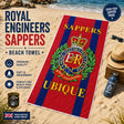 A Royal Engineers Sappers beach towel laid out on a sandy beach next to a straw hat, sunglasses, and a water bottle. The towel features red and navy blue vertical stripes with the Royal Engineers crest in the center and the word 'UBIQUE' printed in yellow at the bottom.