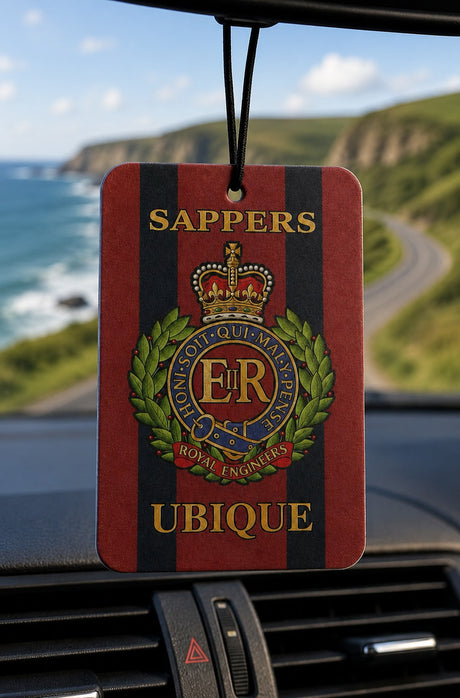 A hanging car air freshener with a red and black striped design, featuring the Royal Engineers Sappers emblem and the words 'SAPPERS UBIQUE'. The product is shown hanging in front of a car dashboard with a blurred background of a coastal road and cliffs.