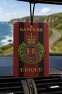A hanging car air freshener with a red and black striped design, featuring the Royal Engineers Sappers emblem and the words 'SAPPERS UBIQUE'. The product is shown hanging in front of a car dashboard with a blurred background of a coastal road and cliffs.