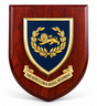 A custom wooden shield plaque featuring a dark blue center shield with a gold lion and laurel wreath, mounted on a reddish-brown wood-grain backing. A gold scroll at the bottom reads 'THE KING'S OWN ROYAL REGIMENT'.